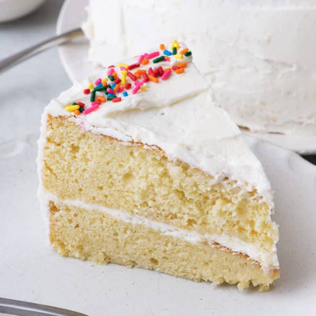 Slice of 2 layer white cake with frosting and sprinkles on a small plate with a fork and whole cake behind it.