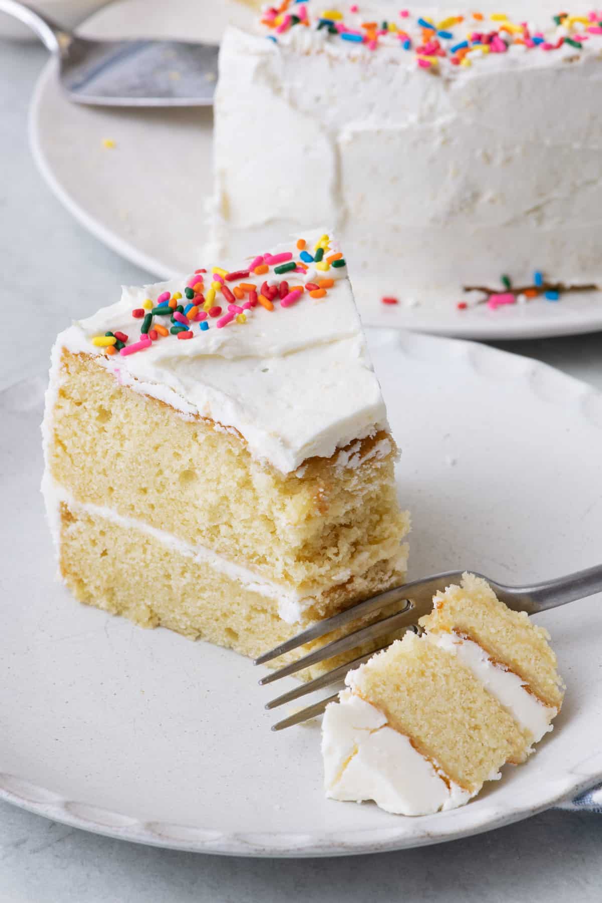 Slice of 2 layer white cake with white frosting and sprinkles with a fork cutting into it.