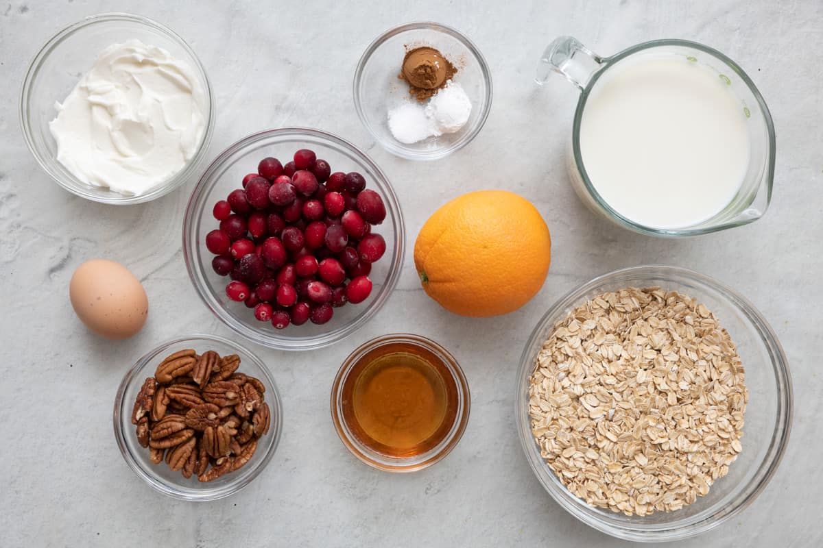 Ingredients for recipes in individual dishes and before prepped: yogurt, 1 whole egg, pecan halves, fresh cranberries, spices, whole orange, maple syrup, milk, and oats.
