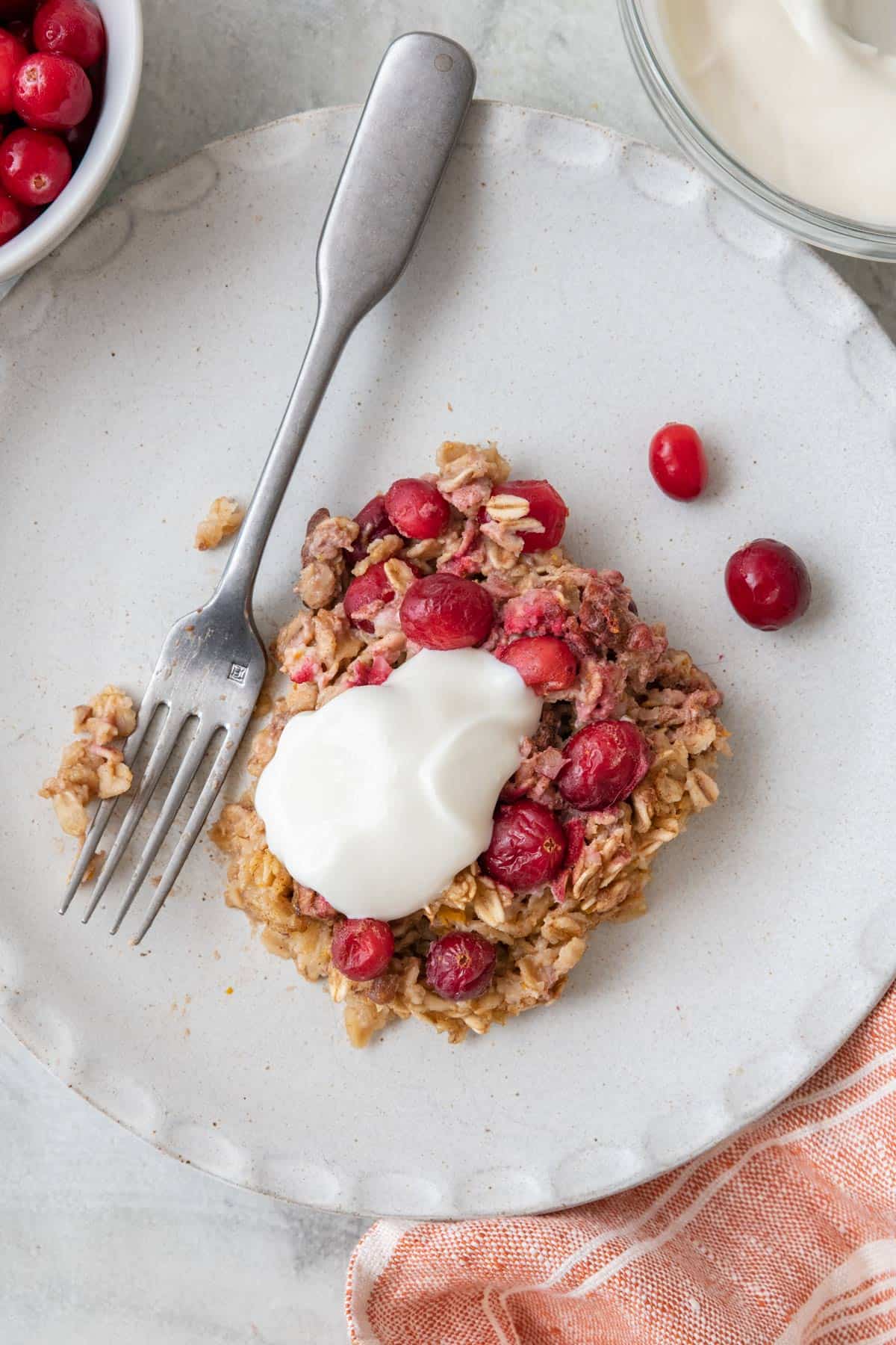 Cranberry pecan baked oatmeal served on a small round plate with a fork and fresh cranberries and yogurt on top and nearby.