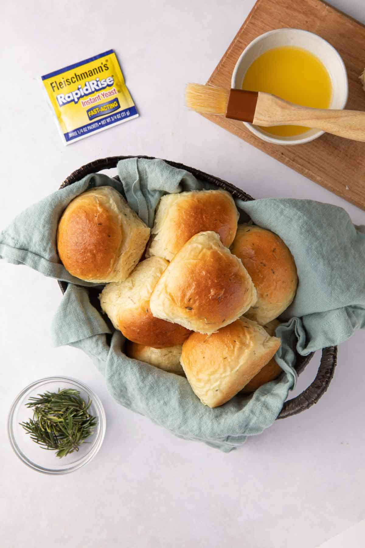 Linen lined oval basket filled with skillet rolls with a hand picking one up, a side dish of fresh rosemary sprigs, a package of Fleischmann's Rapid Rise Instant Yeast, and a small dish of melted butter with a brush resting on top.
