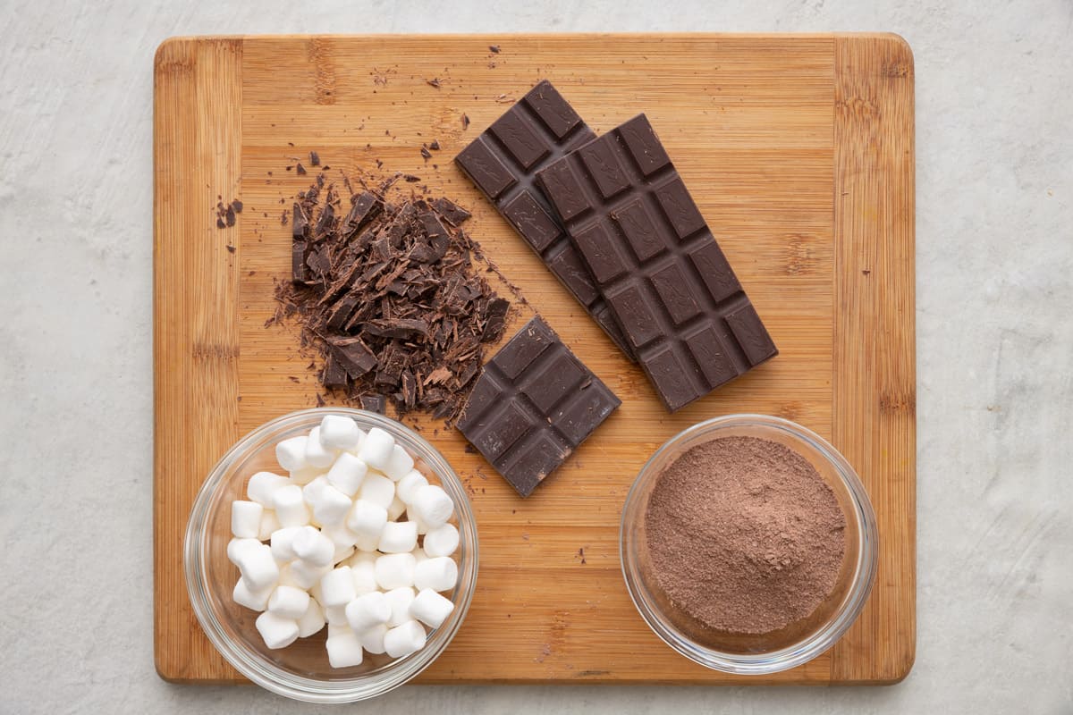 Ingredients for recipe on a cutting board and in individual bowls: mini marshmallows, chocolate bars with one partially chopped, and cocoa powder.