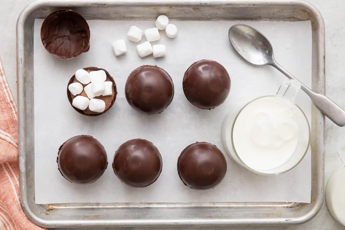 Baking sheet lined with parchment paper with 6 hot chocolate bombs, one opened to show cocoa mix and marshmallows filled inside with a spoon and glass of milk nearby.