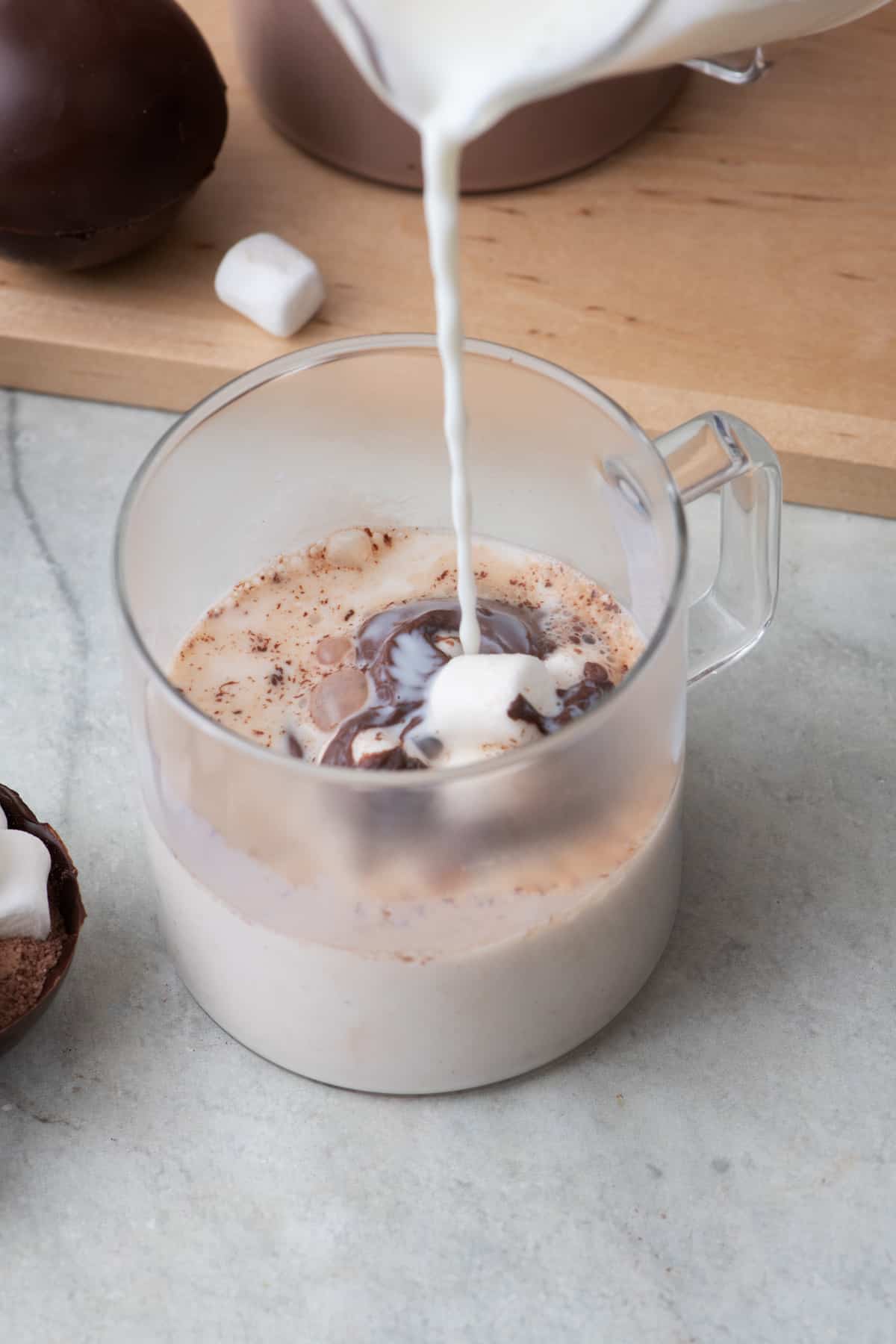Pouring hot milk into a glass mug with a cocoa bomb busting open and melting away to reveal the filling inside.