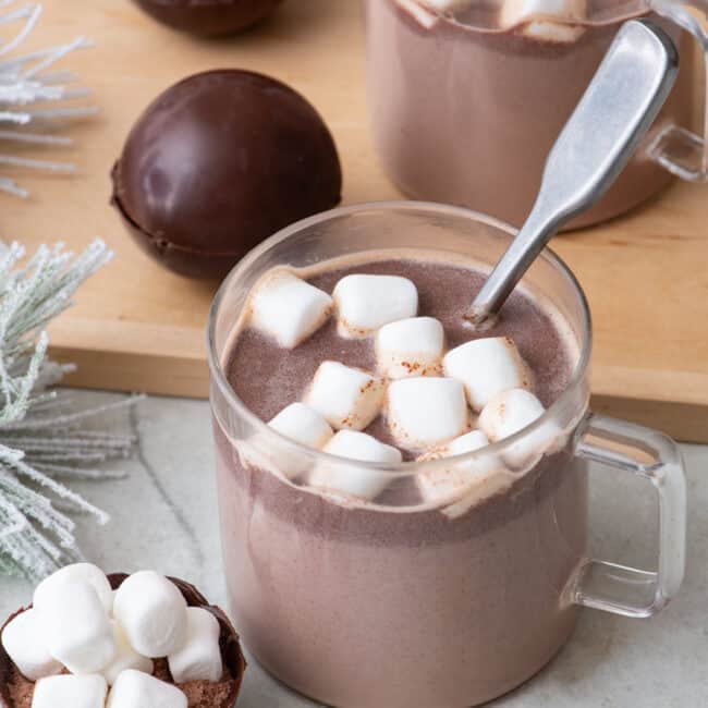 Two glass mugs filled with hot cocoa topped with marshmallows and a spoon dipped in one. Hot cocoa bombs are nearby on a cutting board and one open up front to show inside mix.
