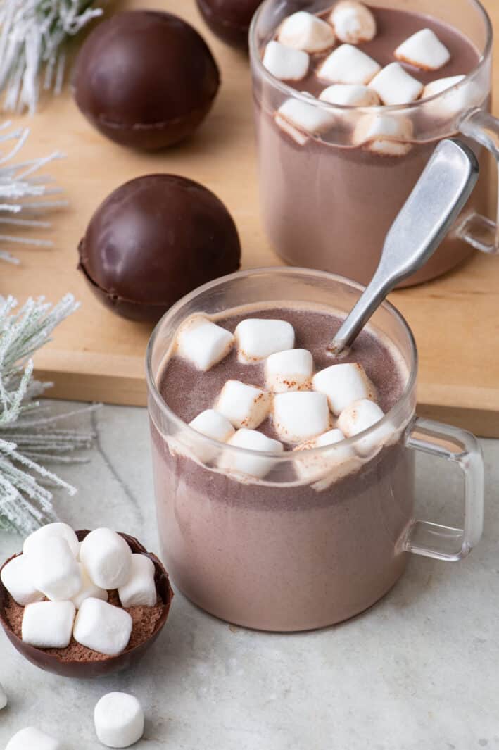 Two glass mugs filled with hot cocoa topped with marshmallows and a spoon dipped in one. Hot cocoa bombs are nearby on a cutting board and one open up front to show inside mix.