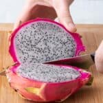 Hand holding half of dragon fruit on cutting board with a knife cut through and other half laying on its side.