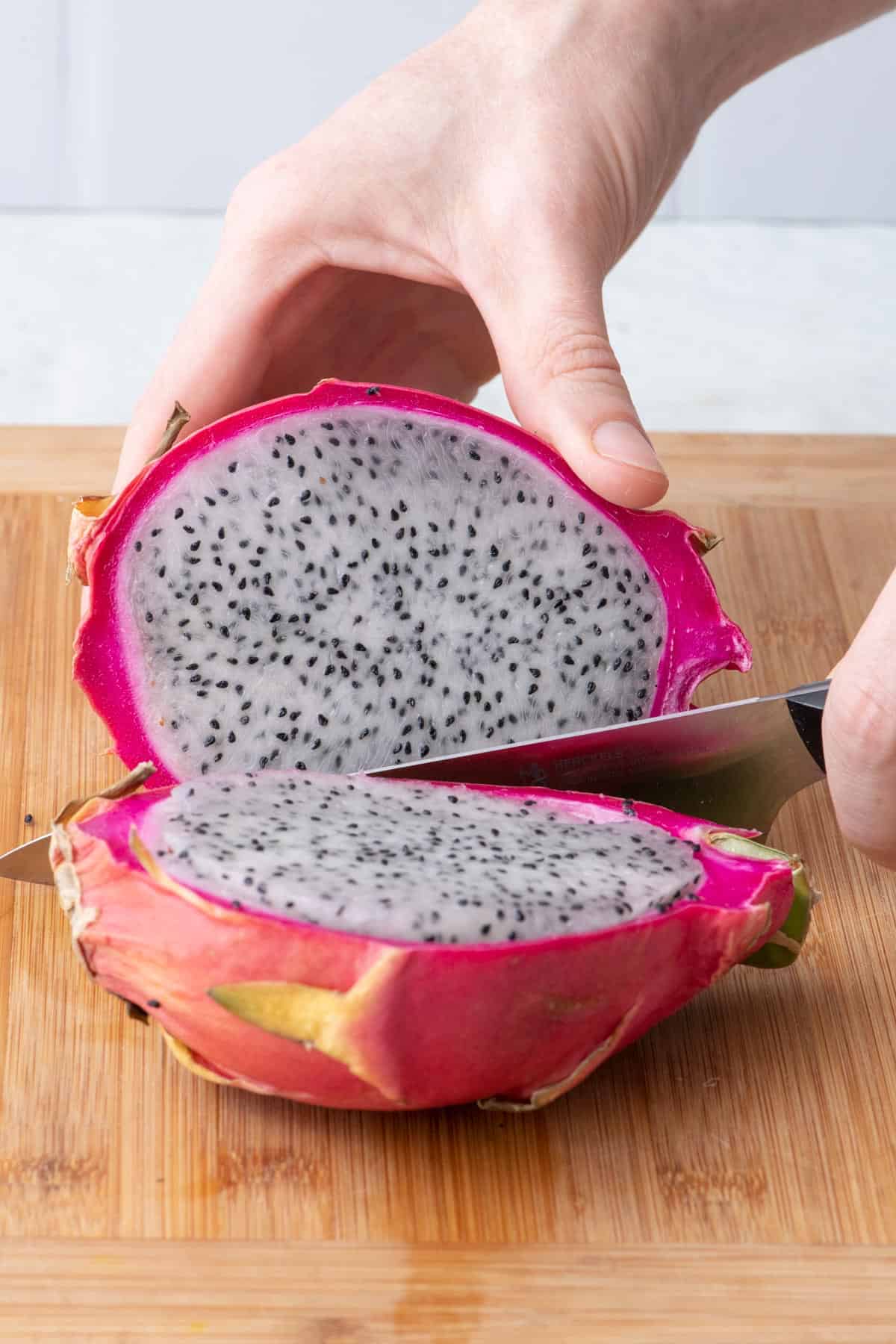 Hand holding half of dragon fruit on cutting board with a knife cut through and other half laying on its side.