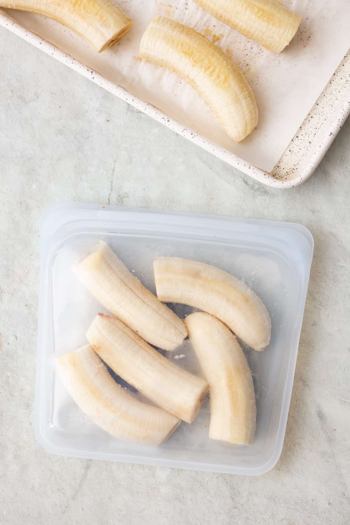 Frozen banana halves inside a reusable freezer bag with tray of them nearby.