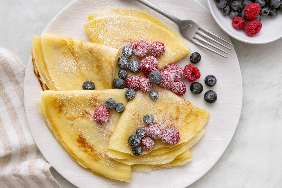 Crepes folded on a white plate topped with fresh berries and dusted with powdered sugar with a fork next to it and a small bowl of more berries nearby.
