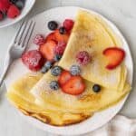 Three delicate crepes folded into trianlges topped with berries and powdered sugar on a small plate with a fork and more berries nearby.