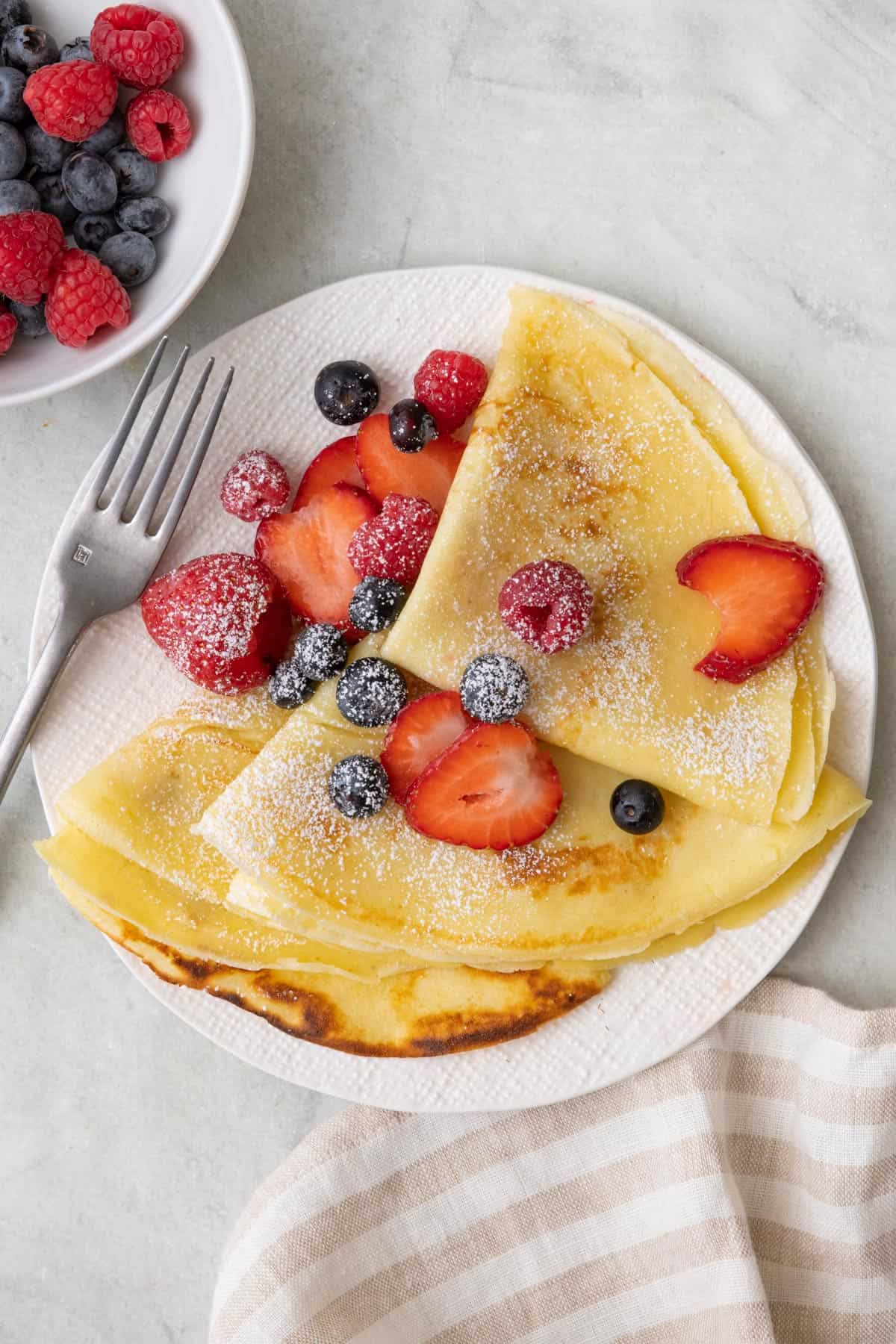 Three delicate crepes folded into trianlges topped with berries and powdered sugar on a small plate with a fork and more berries nearby.