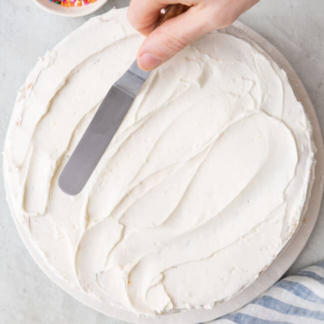 Hand frosting a cake with homemade frosting using a small angled cake spatula with a small dish of sprinkles nearby.