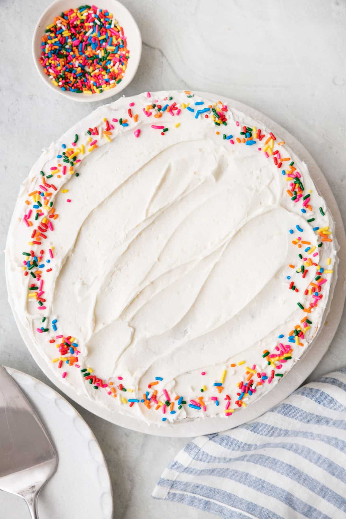 Overhead shot of a frosted cake with sprinles around edge and a small bowl of sprinkles nearby.