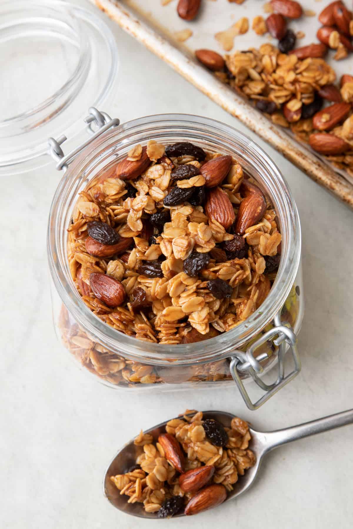 Top down view of homemade granola in an opened small jar with lid, a spoon sitting infront with some on it, and a baking sheet with recipe behind jar.