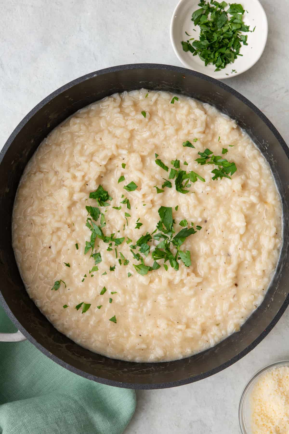 Risotto in pot garnished with fresh parsley with a small dishes of parsley and parmesan nearby.