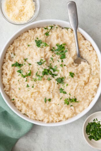 Risotto in serving bowl garnished with fresh chopped parsley and extra parmesan with a spoon dipped in a small dishes of parmesan and parsley nearby.