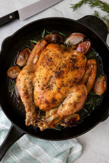 Whole roasted chicken with rosemary and shallots in a cast iron pan with carving knife nearyby.