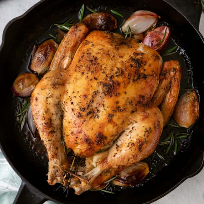 Whole roasted chicken with rosemary and shallots in a cast iron pan with carving knife nearyby.