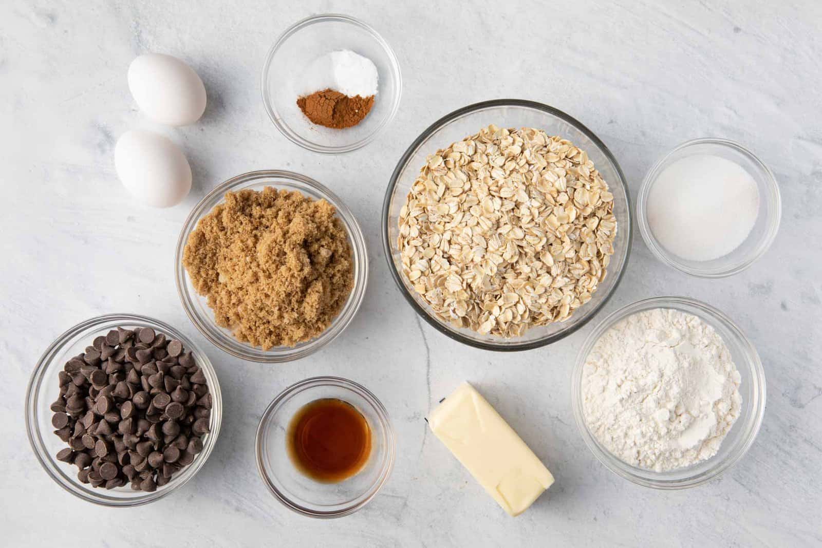 Ingredients for recipe in individual bowls: butter, sugar, oatmeal, chocolate chips, spices, vanilla, flour, 2 eggs, and brown sugar.