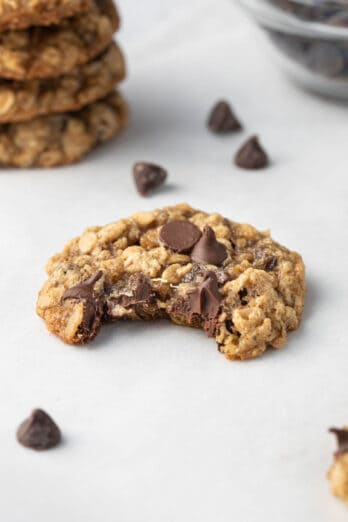 Stack of oatmeal cookies with chocolate chips on parchment paper with one cookie infront with bite taken out and a small dish of chocolate chips in the back.