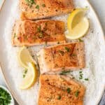 A large oval serving platter with 4 seared salmon filets ontop of a bed of white rice, garnished with lemon wedges and fresh chopped parsley.