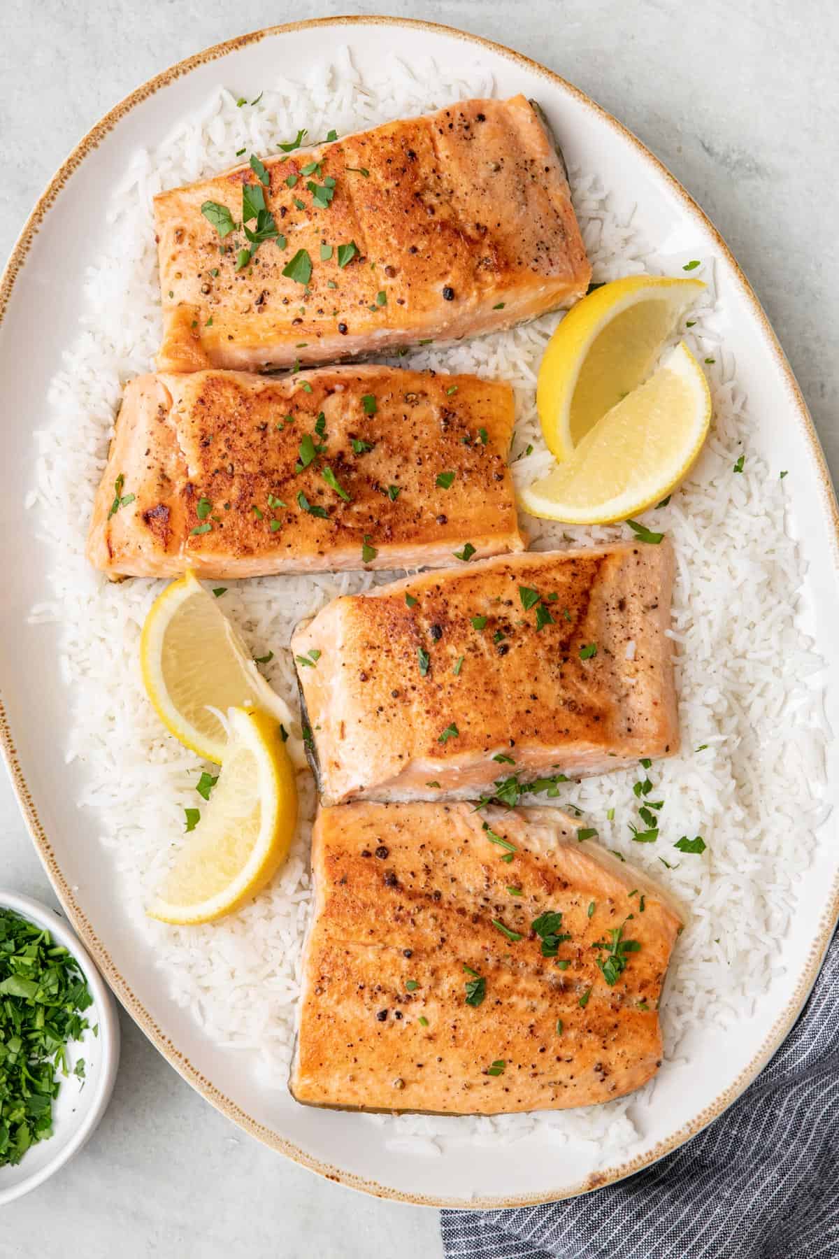 A large oval serving platter with 4 seared salmon filets ontop of a bed of white rice, garnished with lemon wedges and fresh chopped parsley.