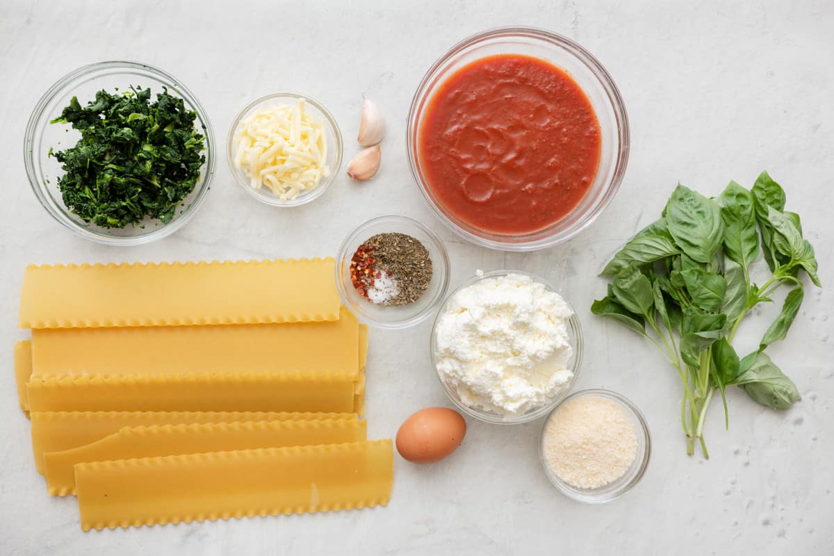 Ingredients for recipe: drained spinach, lasagne noodles, shredded cheese, garlic cloves, seasonings, an egg, red pasta sauce, ricotta, parmesan cheese, and fresh basil.