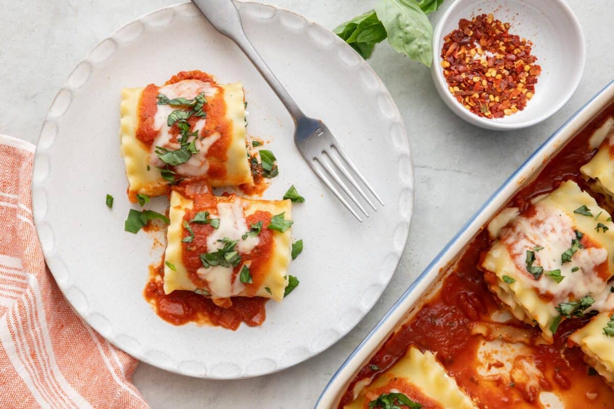 Two lasagne roll ups on a small plate with fork with the baking dish off to side and a small dish of red pepper flakes nearby.