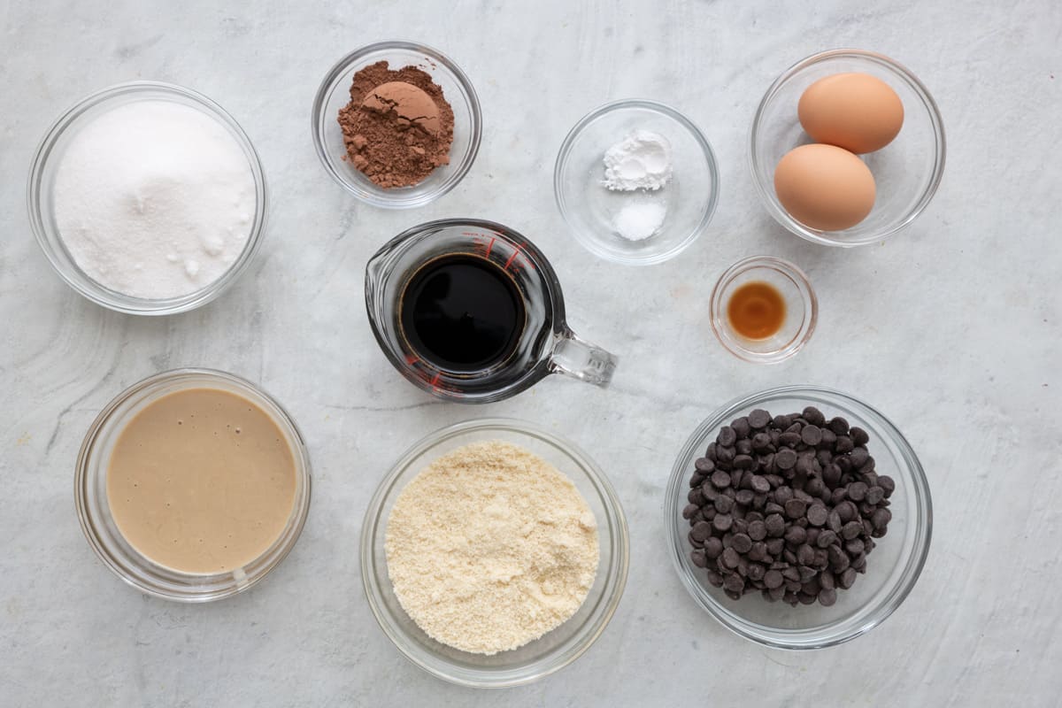 Ingredients for recipe in individual bowls: sugar, tahini, cocoa powder, coffee, almond flour, salt and baking powder, vanilla, 2 eggs, and chocolate chips.