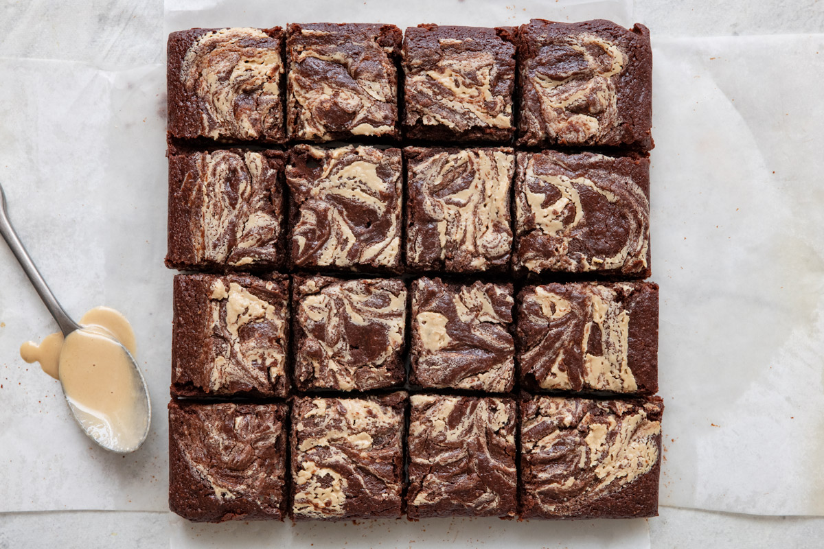 Brownies with tahini swirl cut into 16 squares on parchment paper and a spoon with tahini dripping off nearby.