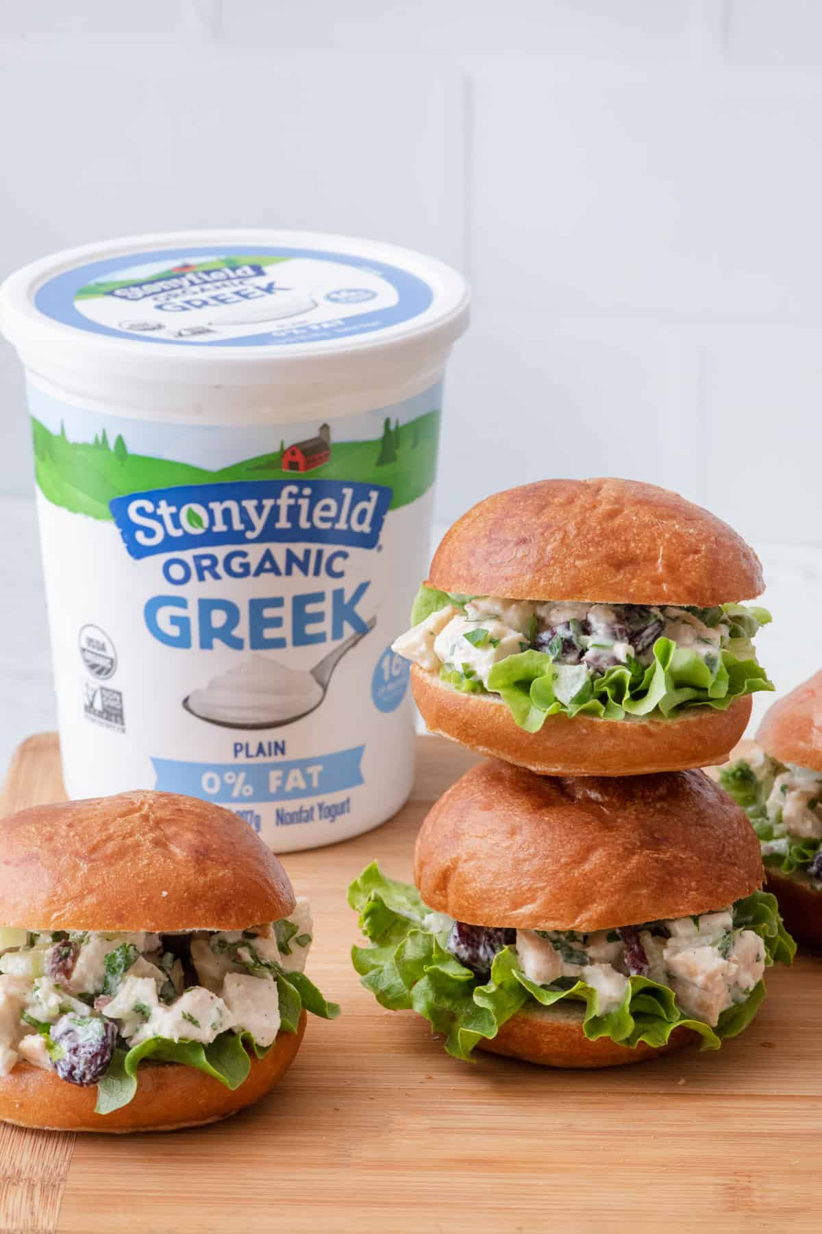 A few turkey salad sandwiches on a cutting board with two stacked on top of eachother and a tub of Stonyfield Organic Greek Yogurt behind them.