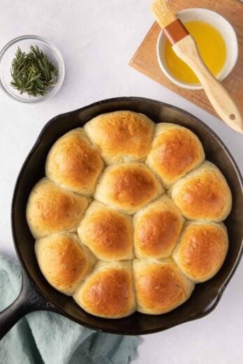 Garlic herb Skillet Dinner Rolls basted with butter in a cast iron skillet with fresh sprigs of rosemary and extra butter and brush nearby.