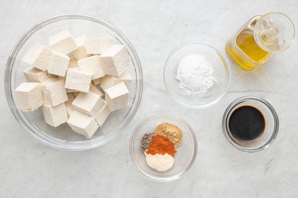 Ingredients for recipe in individual bowls: pressed tofu cubes, cornstarch, seasonings, soy sauce, and oil.