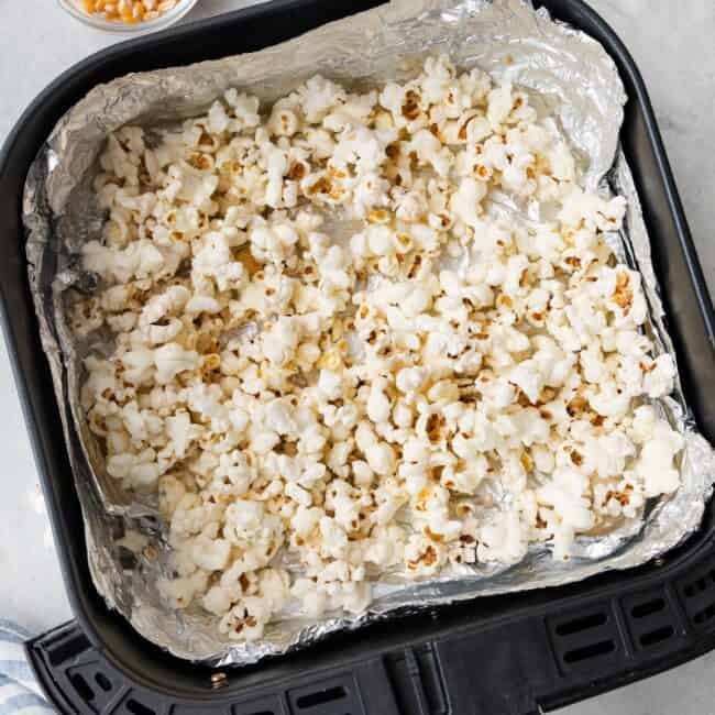 Popcorn in an air fryer basket lined with foil after cooked to show popped kernals with a small dish of uncooked kernals and salt nearby.