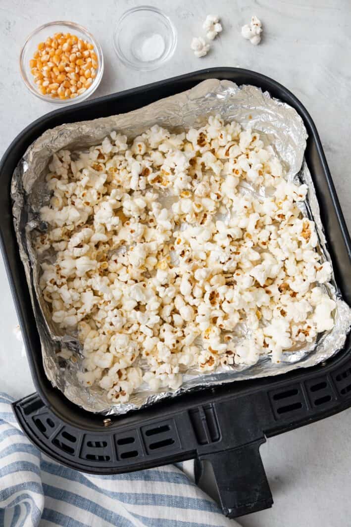 Popcorn in an air fryer basket lined with foil after cooked to show popped kernals with a small dish of uncooked kernals and salt nearby.