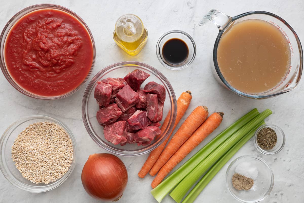 Ingredients for recipe before being prepped: crushed tomatoes, pearl barley, oil, cubed beef, whole onion, Worcestershire sauce, 3 whole carrots, 3 celery stalks, beef stock, Italian seasoning, salt and pepper.