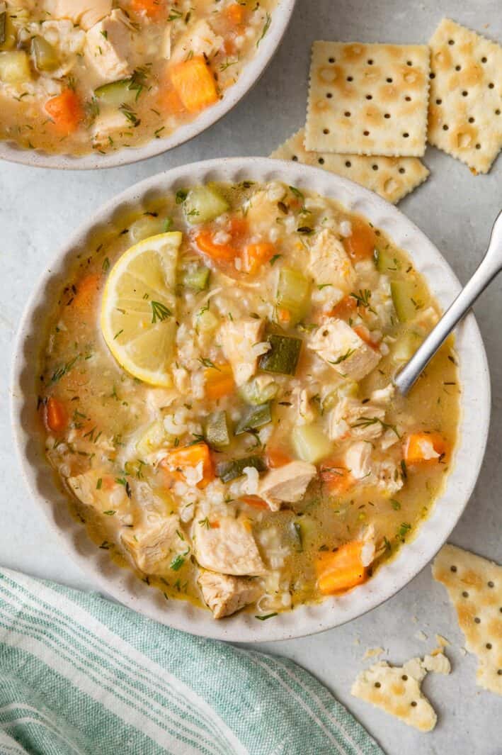 Bowl of chicken vegetable soup garlinished with dill, a spoon dipped inside and crackers nearby.