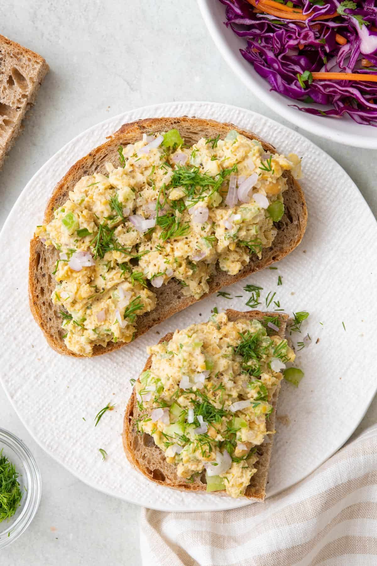 Chickpea "tuna" salad on top of a slice and a half of hearty artisan bread, garnished with dill, with a bowl of red cabbage slaw nearby.