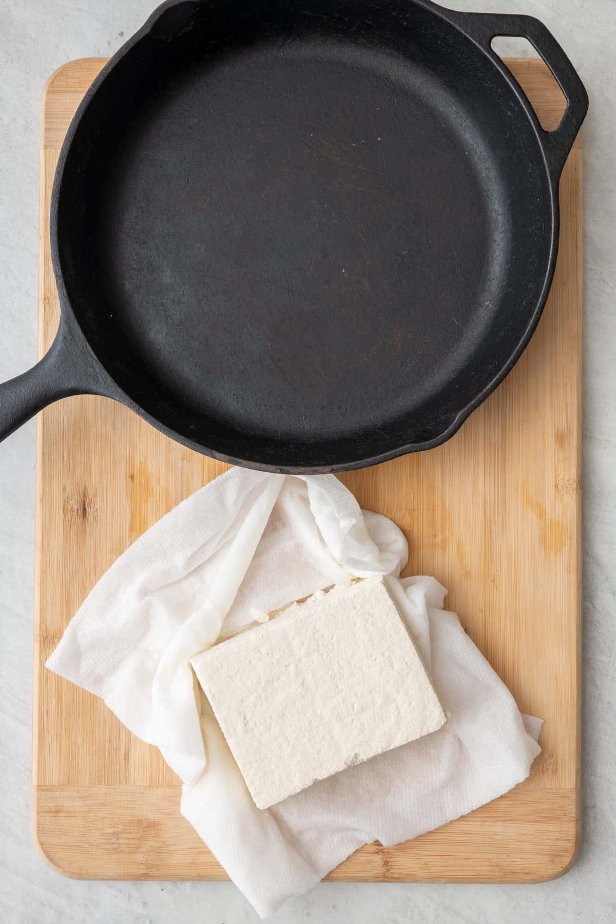 Block of tofu sitting on top of paper towels next to a cast iron skillet on cutting board.