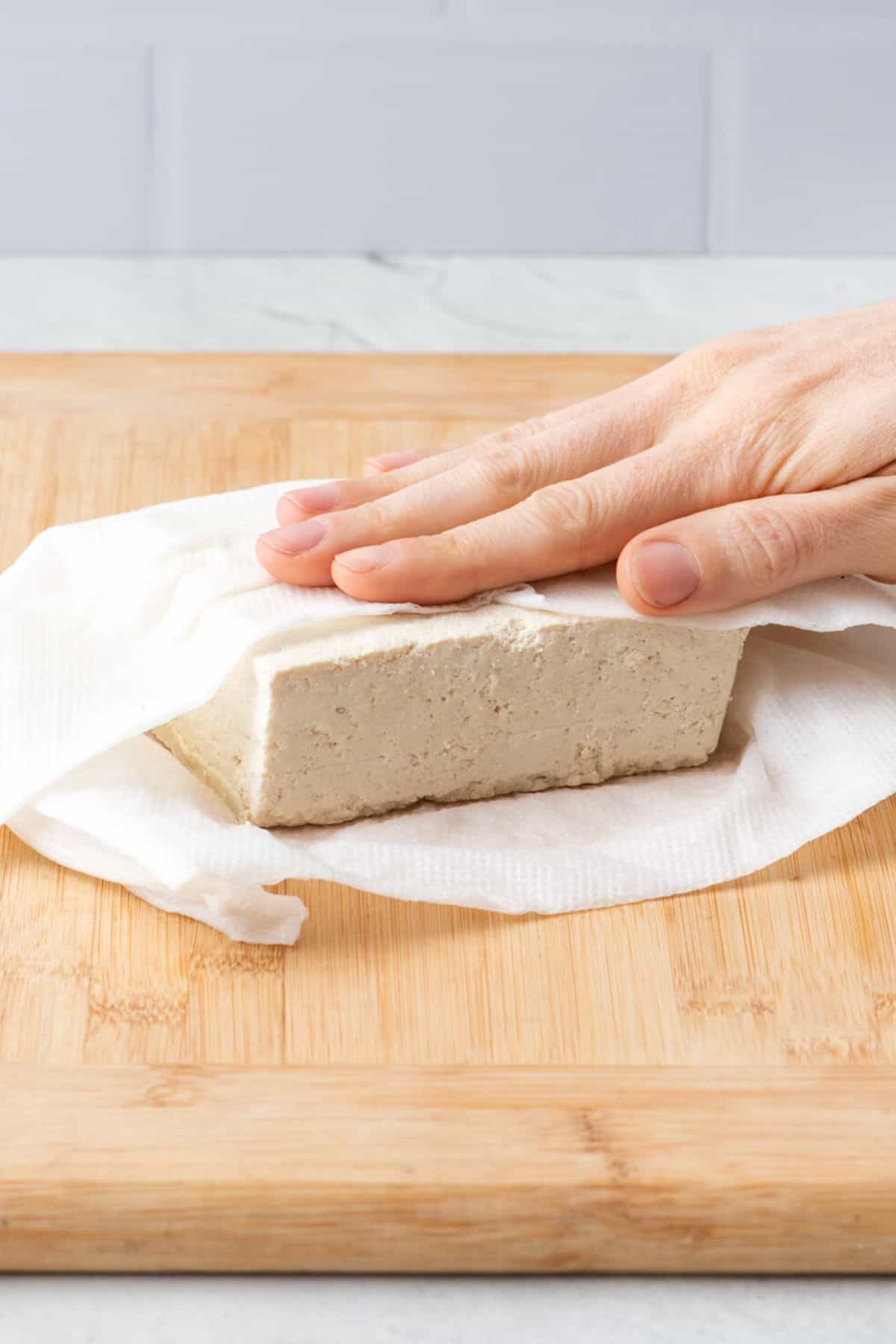 Hand firmly pressing tofu that is wrapped in paper towels on a cutting baord.