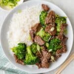 Shallow bowl with rice with beef and broccoli served over, garnished with green onions and seasame seeds with small bowls of extra nearby and a pair of chopsticks.