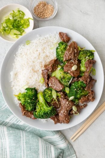 Shallow bowl with rice with beef and broccoli served over, garnished with green onions and seasame seeds with small bowls of extra nearby and a pair of chopsticks.