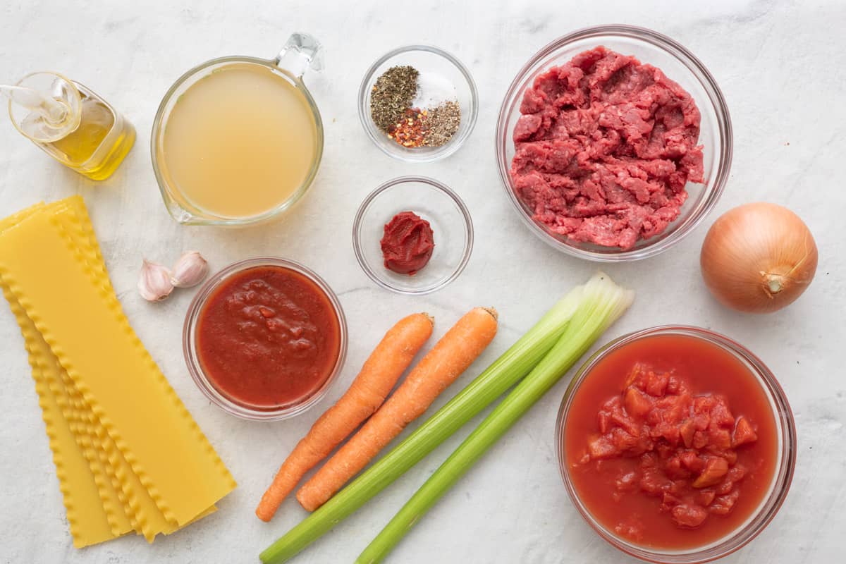 Ingredients for recipe before prepped or in bowls: lasagna noodles, oil, broth, garlic cloves, tomato sauce, seasonings, paste, ground beef, whole carrots, celery stalks, diced tomatoes, and onion.