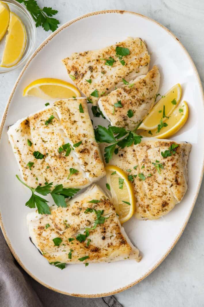4 cooked halibut fillets on oval platter garnished with lemon wedges and fresh chopped parsley.