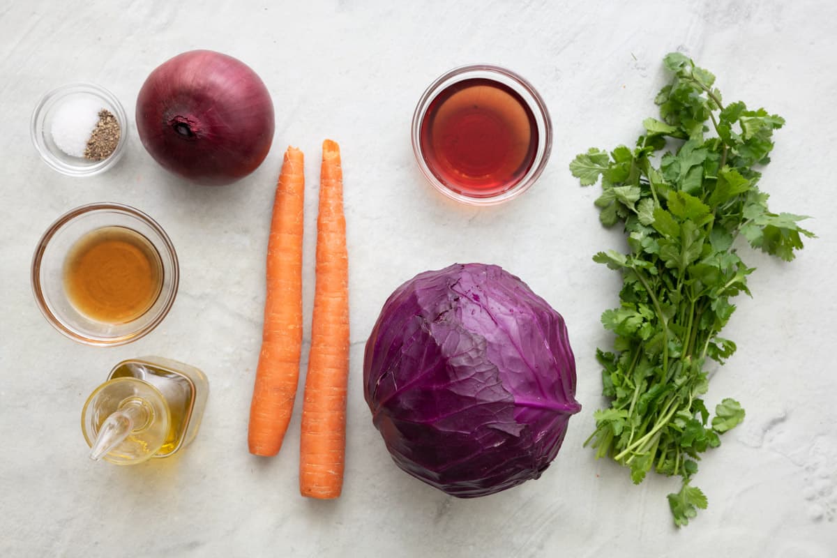 Ingredients for recipe before prepped: salt and pepper, maple syrup, oil, whole red onion, 2 carrots, red wine vinegar, head of red cabbage, and fresh cilantro.