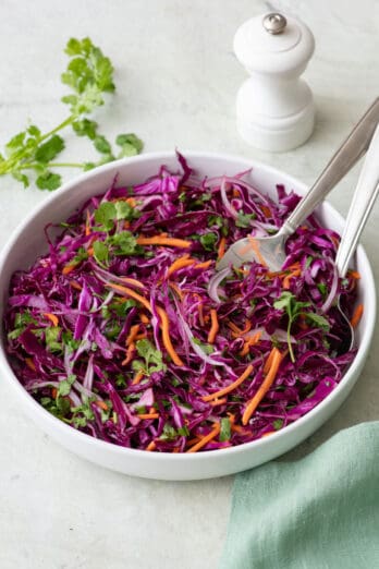 Red cabbage slaw in serving bowl with tongs .