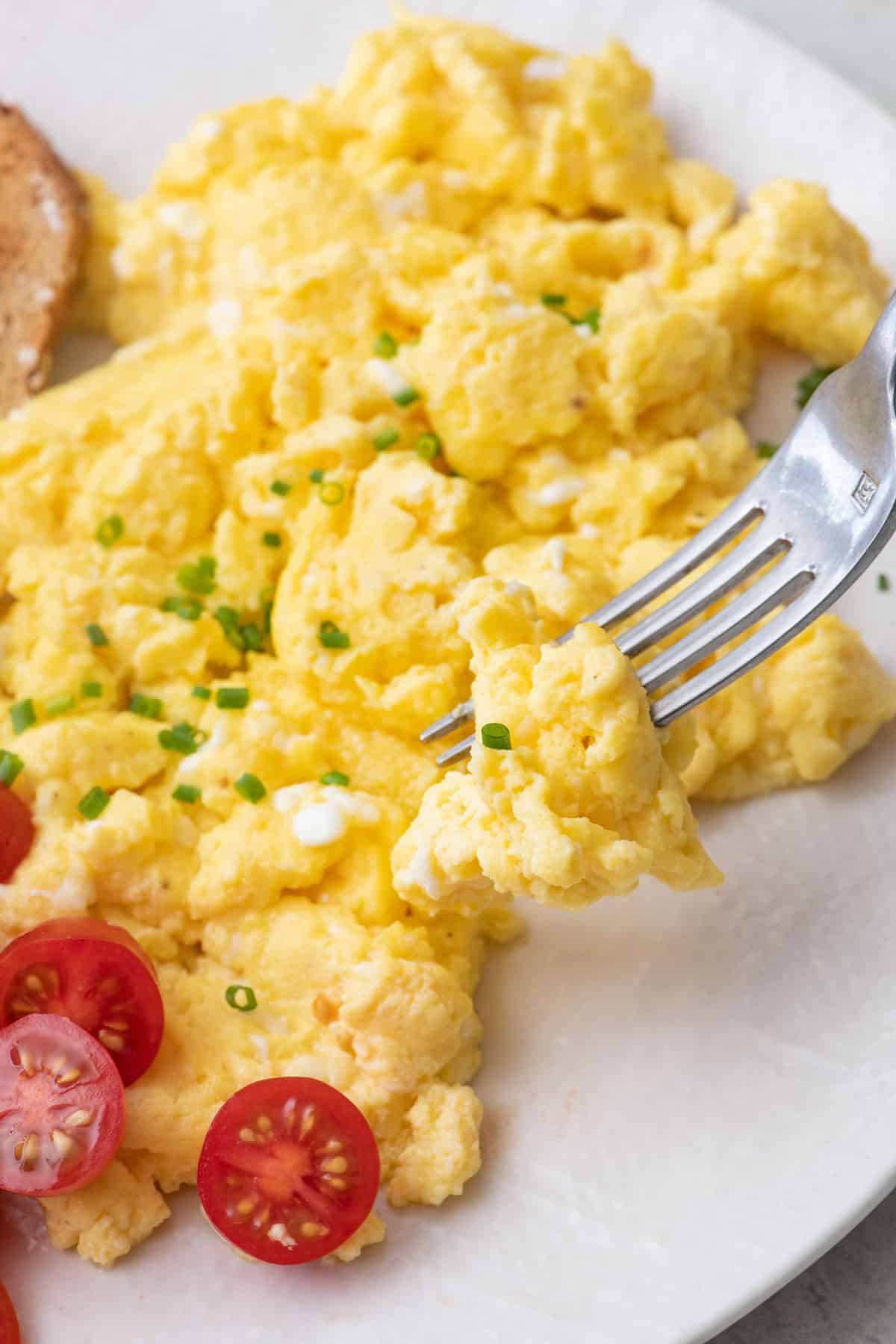 Close up of a fork scooping up some cottage cheese scrambled eggs garnished with chives and a few tomato halves.