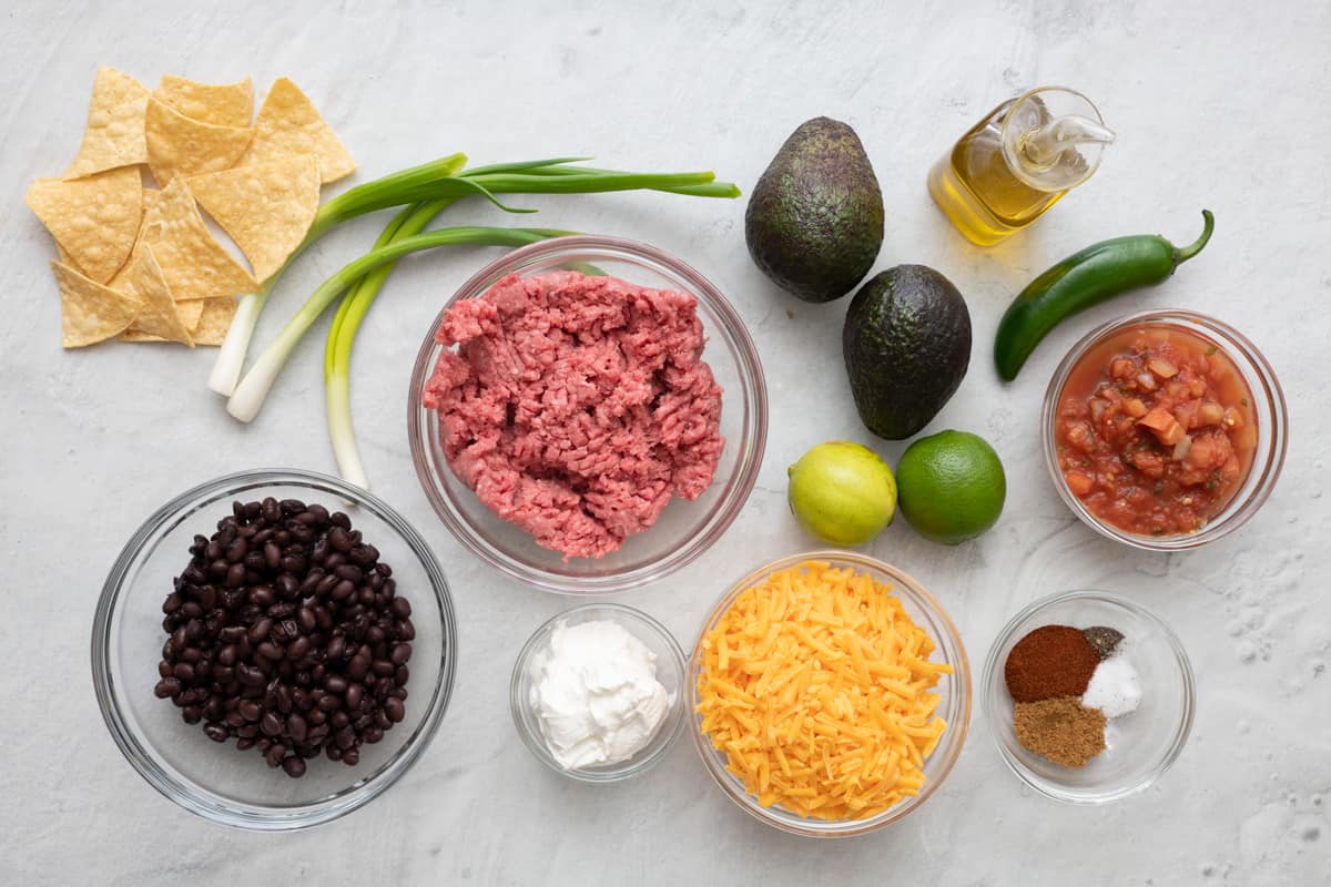 Ingredients for sheet pan recipe: tortilla chips, scallions, black beans, ground beef, sour cream, avocados, limes, shredded cheese, oil, jalepeno, salsa, and spices.