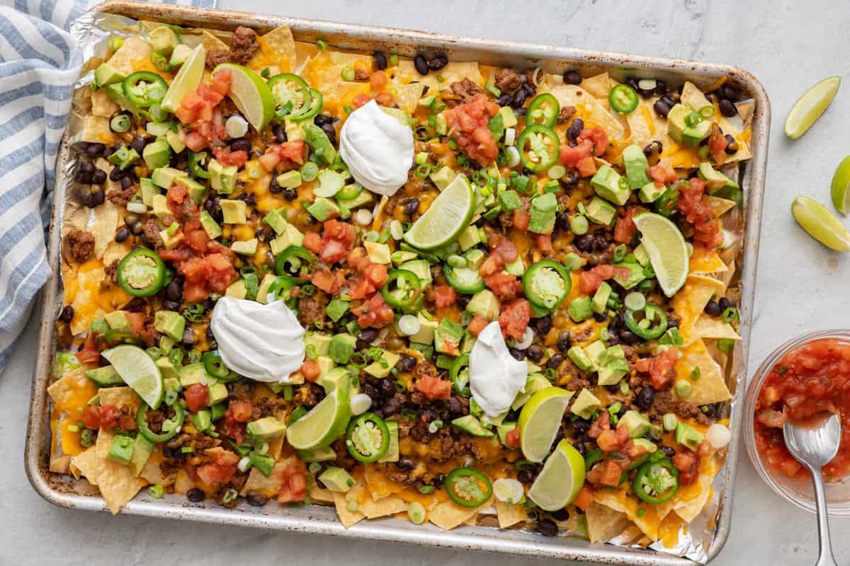 Baking tray of nachos with veggie toppings, a dollop of sour cream and salsa, and a small dish of salsa nearby with spoon dipped in.